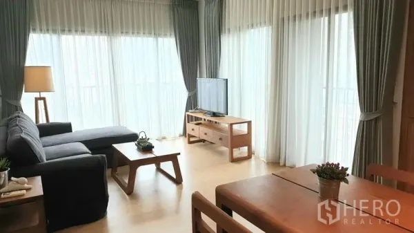 Condominium for rent or sale in Watthana, Bangkok - Corner living space with TV console and wraparound windows dressed in sheer drapes.