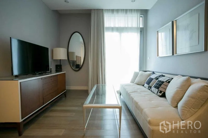 คอนโด ให้เช่าในPathum Wan, Bangkok - มุมนั่งดูทีวี อโซฟาทรงยาว โต๊ะกลางกระจก และกระจกเงาทรงวงรีข้างหน้าต่าง