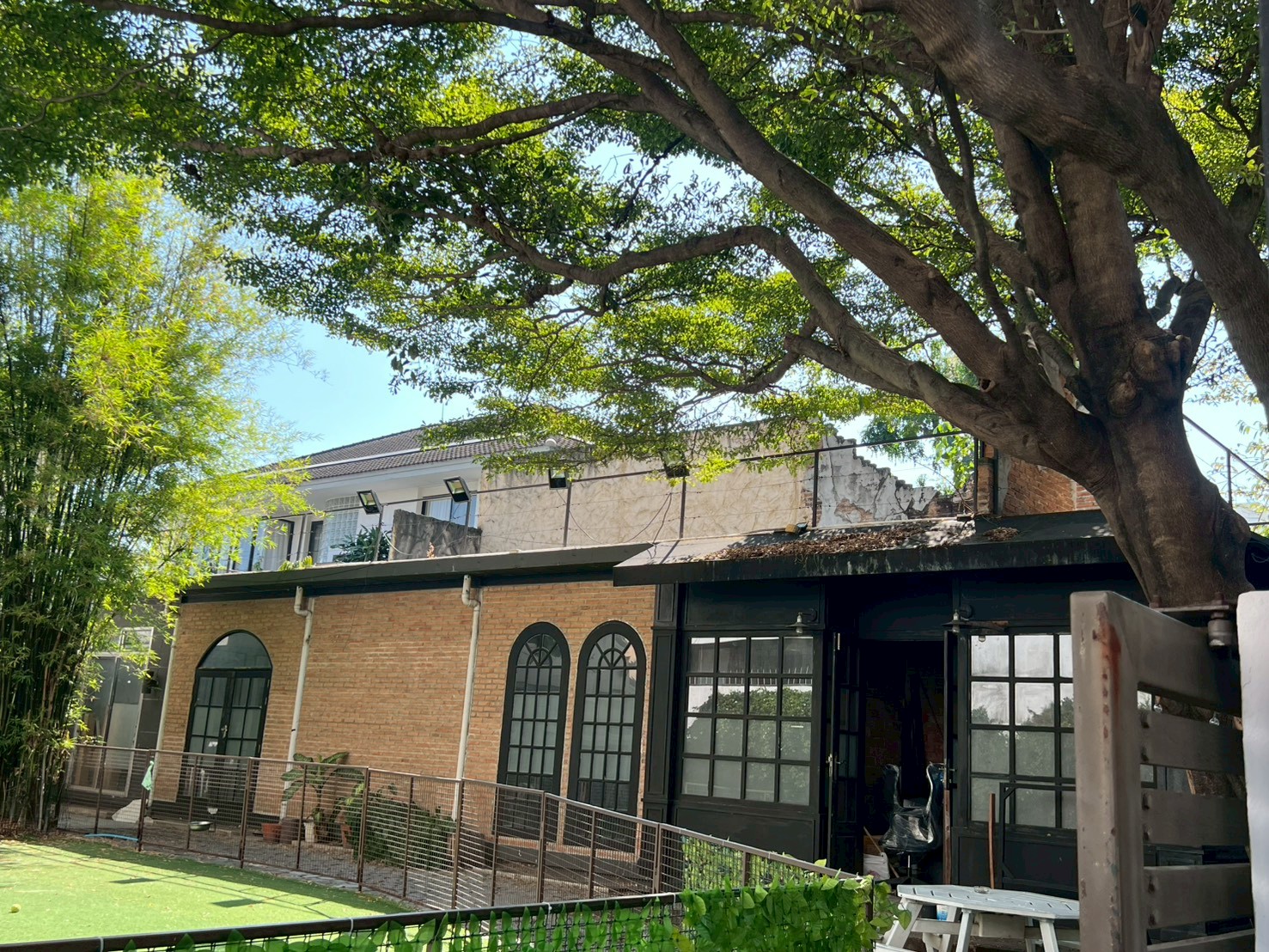 零售空间 出租在Watthana, Bangkok - 绿荫庭院与大树，面向带拱窗的砖结构建筑。