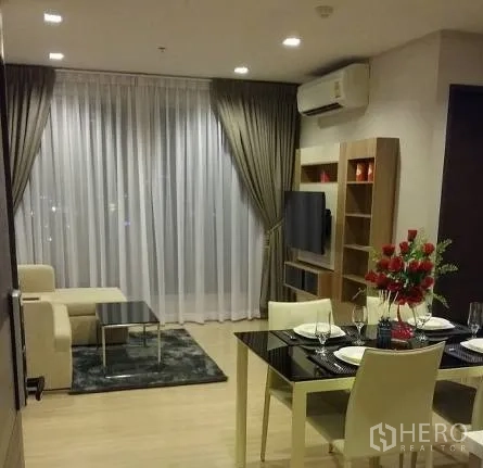 Condominium for rent in Sathon, Bangkok - Open-plan living and dining area with TV unit, four-seat dining table and large windows with drapes.