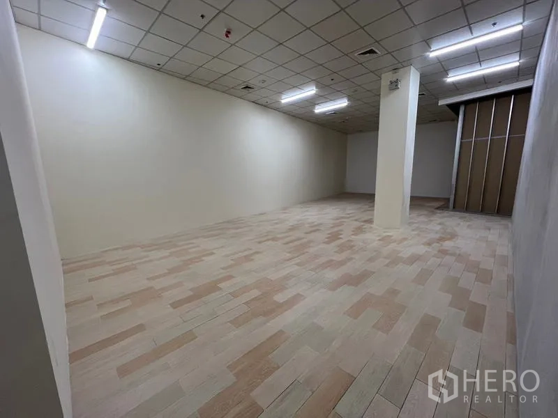 零售空间 出租在Watthana, Bangkok - 空置开间商铺，木纹地砖、网格吊顶、中央立柱与明亮灯带。