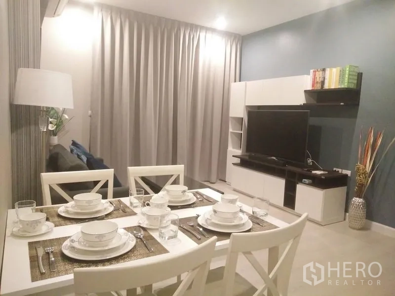 Condominium for rent in Huai Khwang, Bangkok - Bright living and dining area with set table, TV wall unit, and floor-to-ceiling curtains in a modern Bangkok condo.
