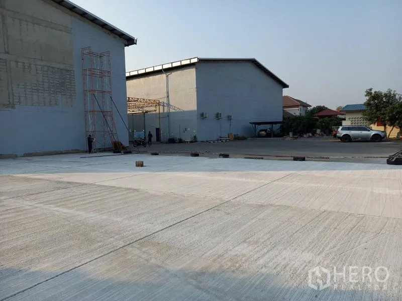 Warehouse for rent in Mueang Pathum Thani, Pathum Thani - Open concrete yard beside warehouse buildings with scaffolding and parking area, suitable for loading and maneuvering.