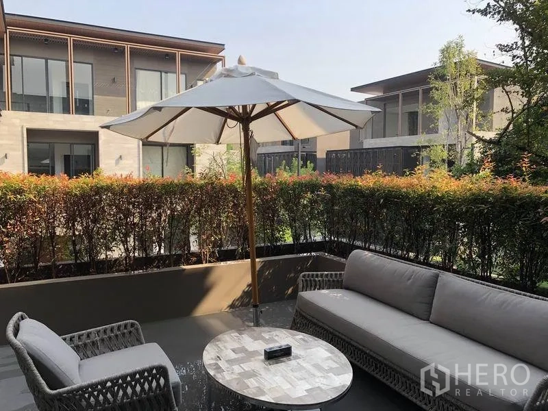独立式住宅 出售在Bang Kapi, Bangkok - 私家露台配遮阳伞与藤编座椅，修剪整齐的绿篱与现代邻里景观。