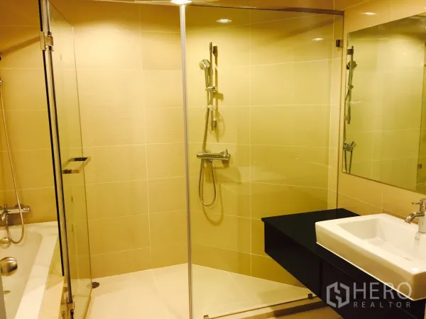 Condominium for rent in Huai Khwang, Bangkok - Modern bathroom with glass-enclosed shower, separate bathtub, large mirror and vanity in neutral tiles.