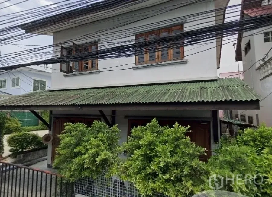 独立式住宅 出售在Phra Khanong, Bangkok - 帕卡侬两层独栋住宅，绿色遮雨棚屋顶、木框窗、带栅栏前庭与路旁电线。