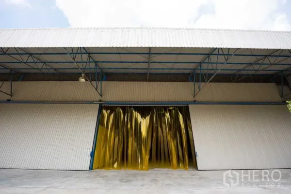 Warehouse for rent in Phra Phutthabat, Saraburi - Wide steel warehouse facade with sliding doors and yellow PVC strip curtain entrance.