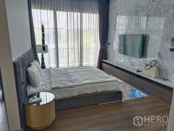 Detached House for rent in Bang Phli, Samut Prakan - Primary bedroom with upholstered bed, marble accent wall with mounted TV and balcony-height windows.