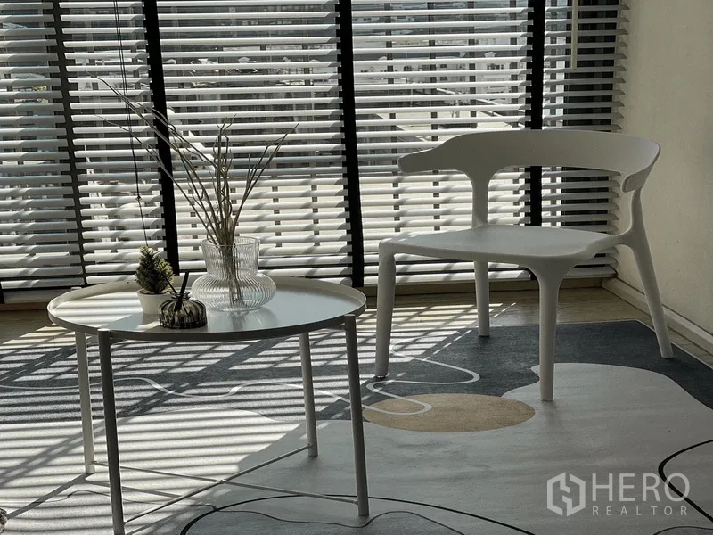 公寓 出租在Khlong Toei, Bangkok - 靠窗百叶旁的白色椅子与圆形茶几特写，铺有图案地毯。