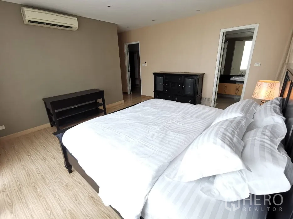 Condominium for rent in Watthana, Bangkok - Master bedroom angle showing ensuite entry, dresser, air conditioning, bedside lamp, and wood floors.