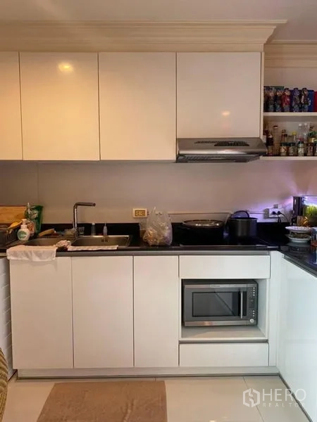 Condominium for rent in Watthana, Bangkok - Compact L-shaped kitchen featuring white cabinetry, black countertop, microwave and cooktop.