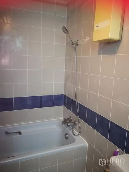 Townhouse for rent or sale in Watthana, Bangkok - Bathroom with bathtub, handheld shower and beige tiles with blue accent strip.