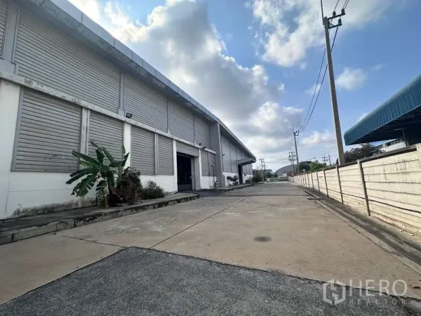 工厂 出租 - 大型工业仓库外立面，卷闸门与宽阔装卸车道，混凝土路面与多云天空。