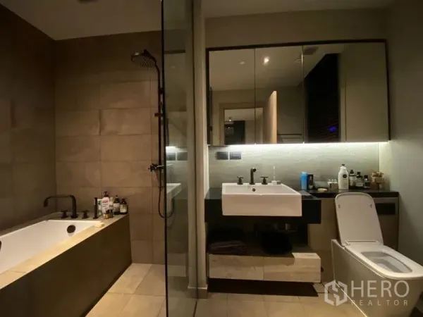 Condominium for rent or sale in Watthana, Bangkok - Contemporary bathroom with soaking bathtub, separate glass shower, large mirror, and toilet in neutral tiles.