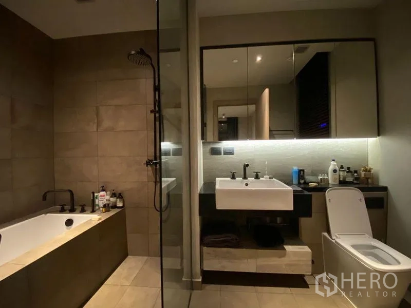 Condominium for rent or sale in Watthana, Bangkok - Contemporary bathroom with soaking bathtub, separate glass shower, large mirror, and toilet in neutral tiles.