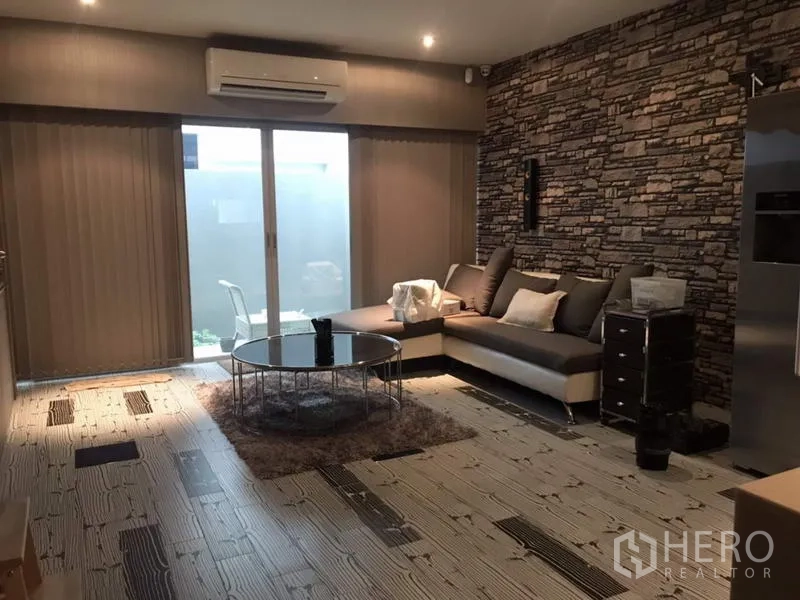 Townhouse for rent or sale in Suan Luang, Bangkok - Modern living room with stone accent wall, L-shaped sofa, round glass coffee table, and sliding doors to a small patio.