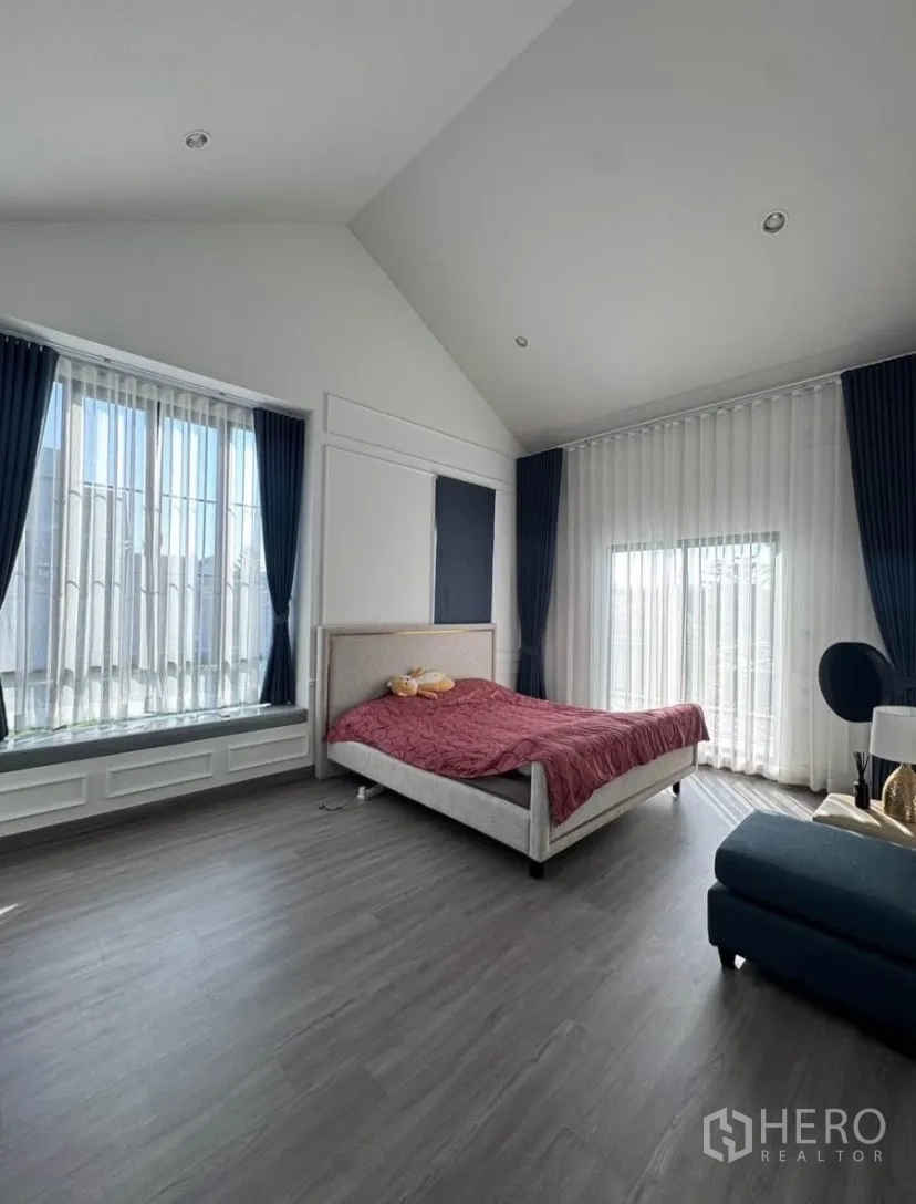 บ้านเดี่ยว for rent or sale in Lat Krabang, Bangkok - ห้องนอนกว้าง เพดานสูงทรงจั่ว หน้าต่างสูงจรดฝ้า ม่านโปร่ง และเตียงบนพื้นลายไม้สีเทา