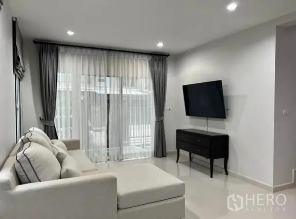 独立式住宅 出租在Suan Luang, Bangkok - 采光充足的客厅，转角沙发、壁挂电视与落地窗帘