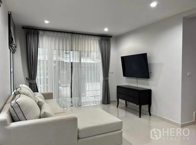 独立式住宅 出租在Suan Luang, Bangkok - 采光充足的客厅，转角沙发、壁挂电视与落地窗帘