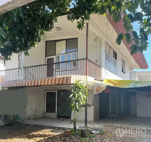 Detached House for sale in Khlong Toei, Bangkok - Shaded front yard and carport beside a two-storey house with balcony and striped awning.