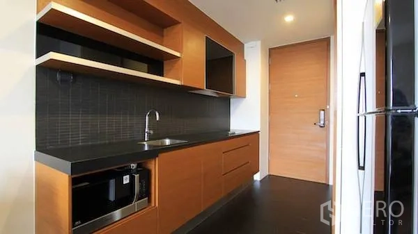 Condominium for rent in Khlong Toei, Bangkok - Modern galley kitchen with wooden cabinets, black countertop, built-in microwave and sink.