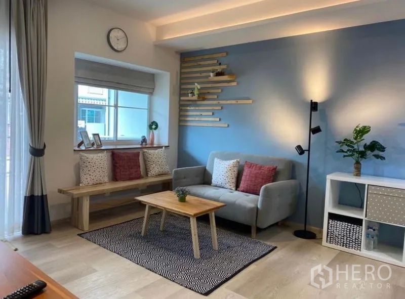 ทาวน์เฮาส์ ให้เช่าในBang Phli, Samut Prakan - ห้องนั่งเล่นสว่างของทาวน์เฮ้าส์บางพลี ผนังสีฟ้า ม้านั่งข้างหน้าต่าง โซฟาสีเทา และโต๊ะไม้เล็ก