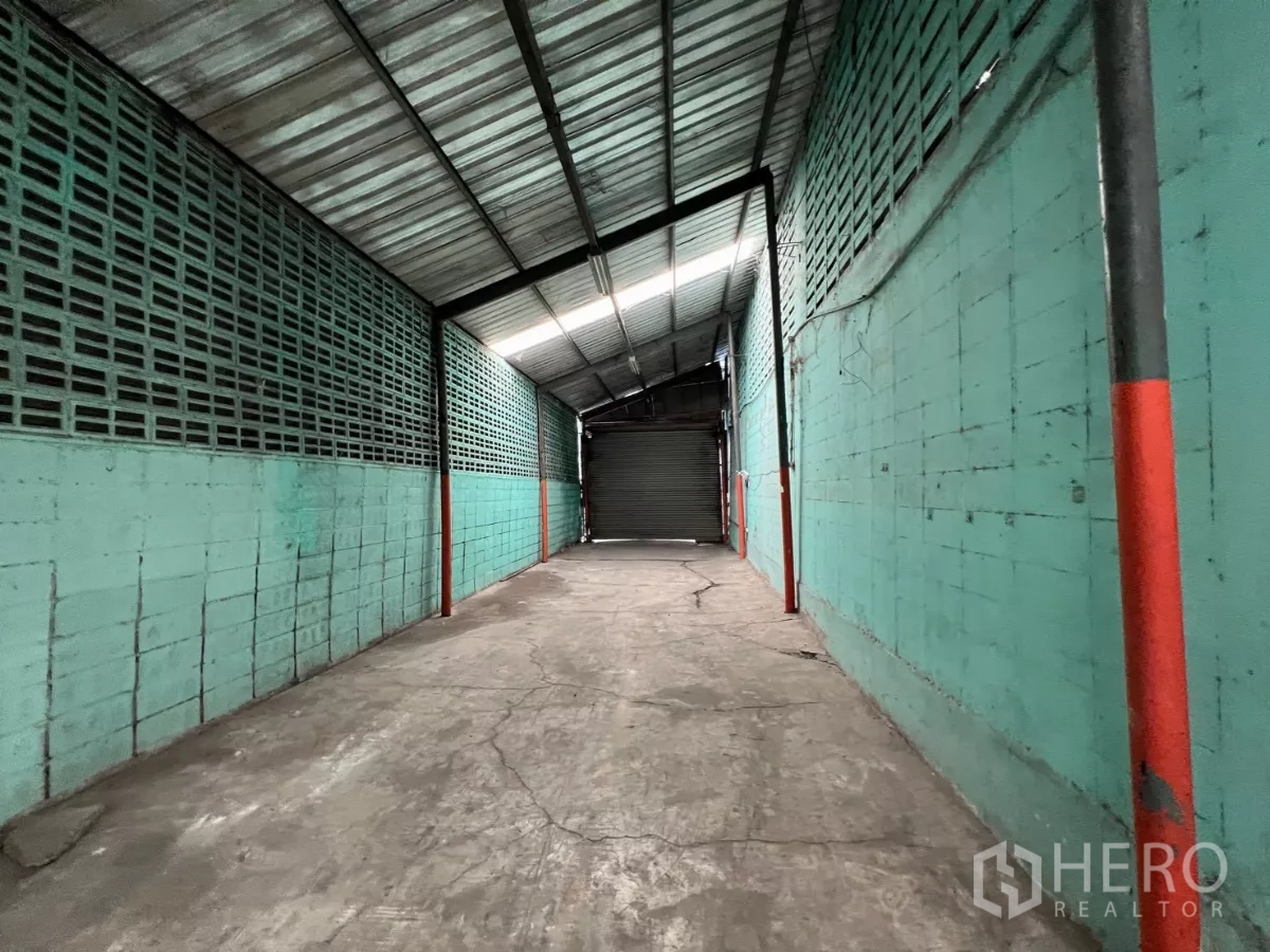 Warehouse for rent in Khlong Sam Wa, Bangkok - Teal block-walled corridor under a metal roof leading toward a roller shutter entry.