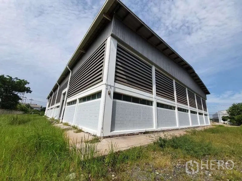 Factory for rent in Mueang Samut Prakan, Samut Prakan - Corner view of a large steel-frame factory with louvered ventilation panels, concrete base and grass verge under a blue sky.