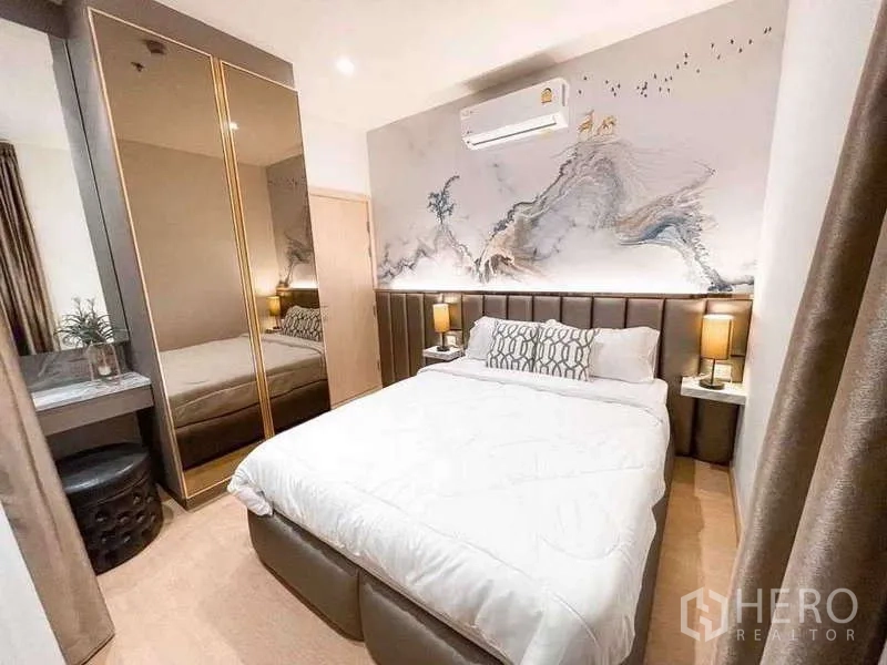 Condominium for rent or sale in Watthana, Bangkok - Warm bedroom with padded headboard, artistic wall panel, mirrored wardrobe, and bedside lamps.