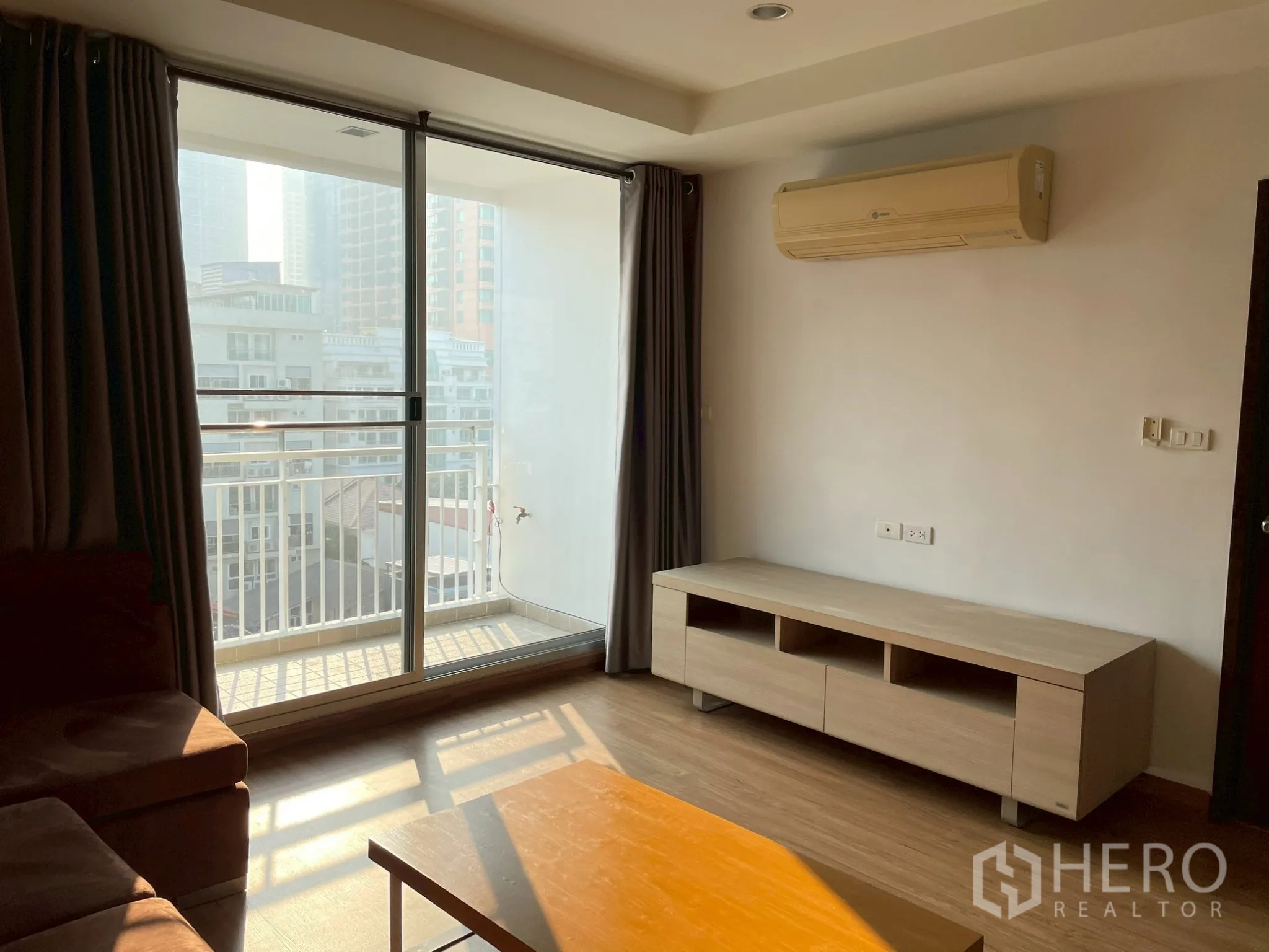 Apartment for rent in Khlong Toei, Bangkok - Living room with sliding balcony doors, wall-mounted air conditioner, and TV console.