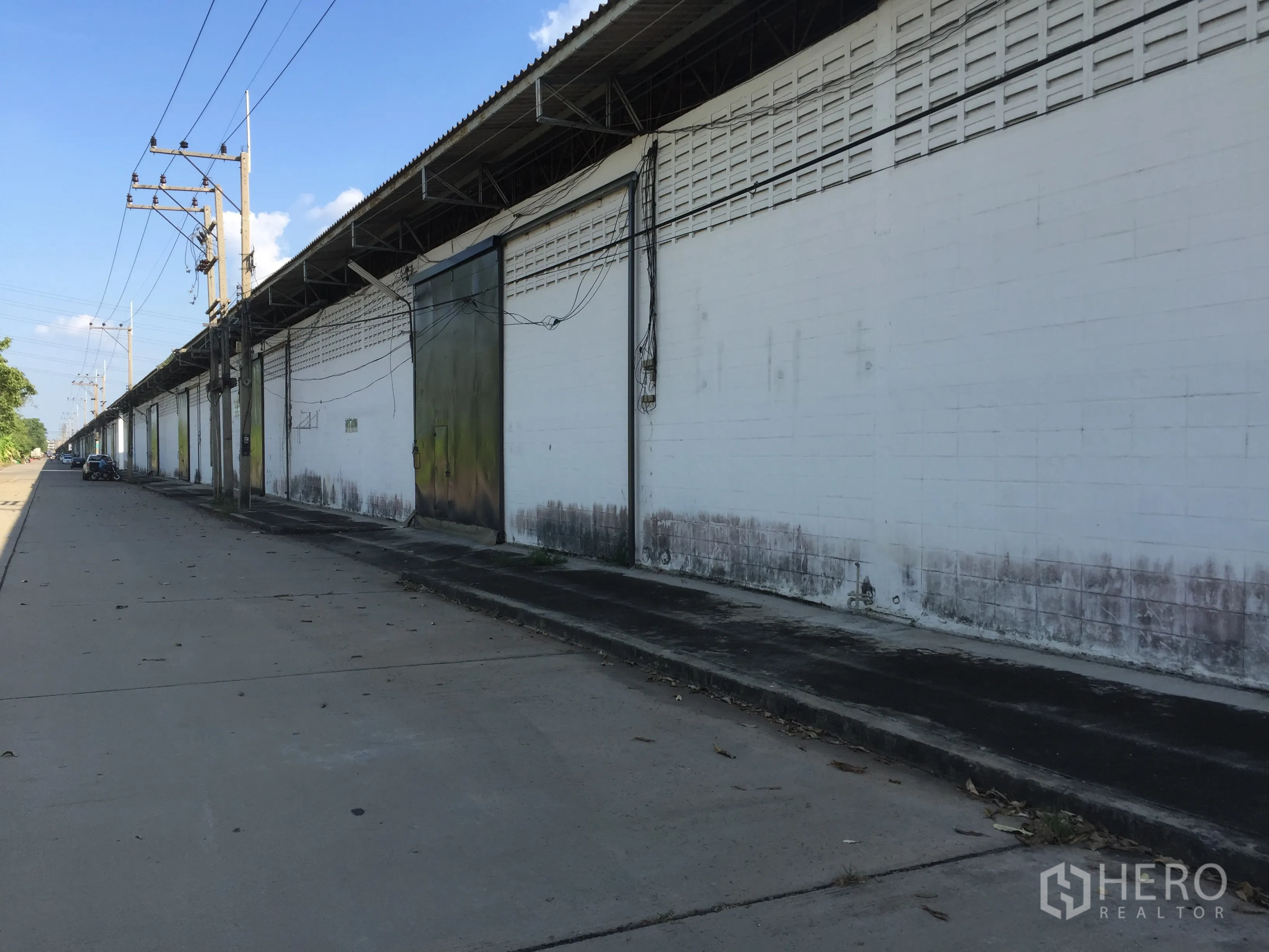 工厂 出租在Pathum Thani - 白色厂房立面沿宽阔混凝土道路延伸，配有电线杆与装卸通道。