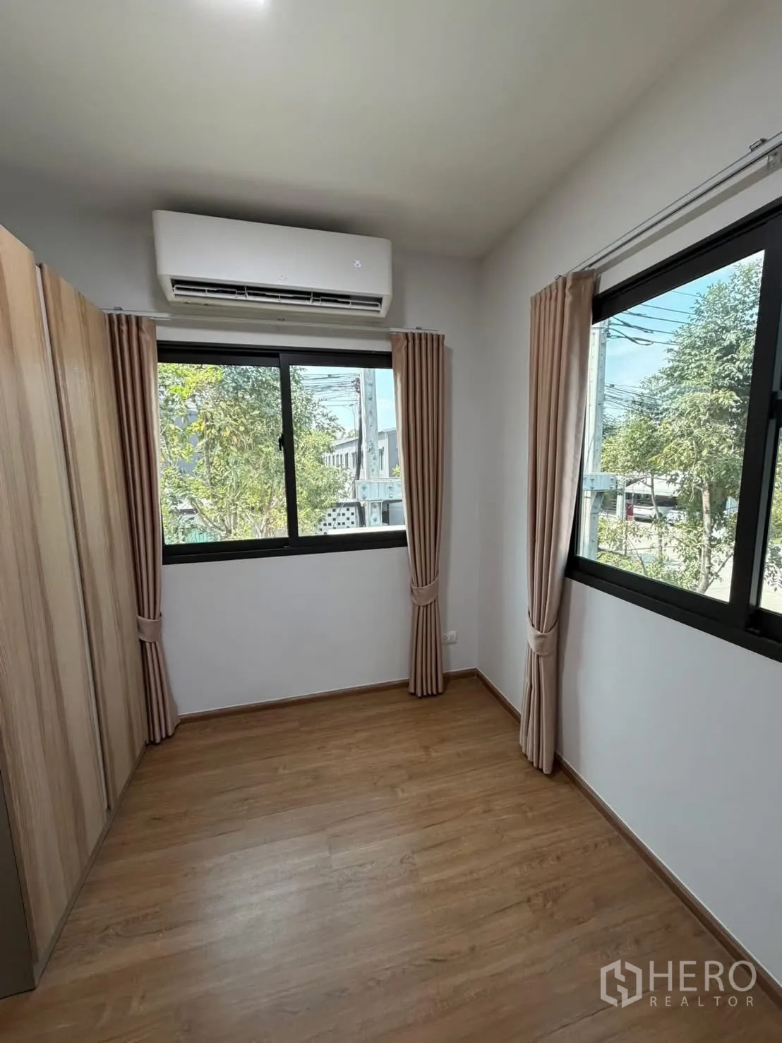 Townhouse for rent in Bang Phli, Samut Prakan - Bright secondary bedroom with two corner windows, air-conditioner, wood floor and neutral curtains.