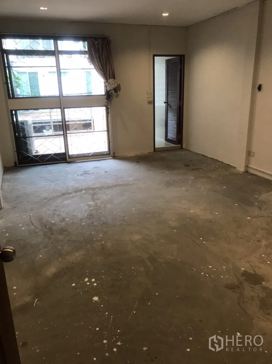 Home Office for rent in Khlong Toei, Bangkok - Unfurnished bedroom with large window and security grilles, raw concrete floor, and a doorway to an ensuite.