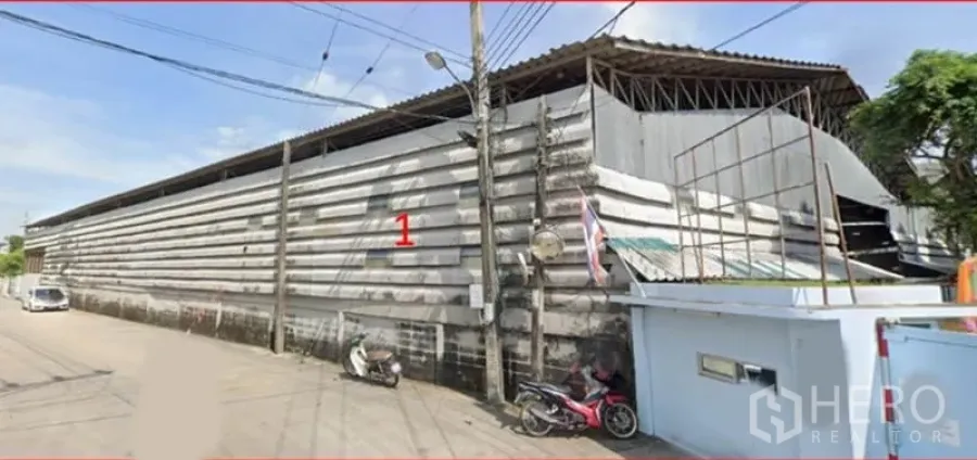 Warehouse for rent in Bang Khun Thian, Bangkok - Street view of a long corrugated-metal warehouse exterior with wide roofline and gated side entrance.