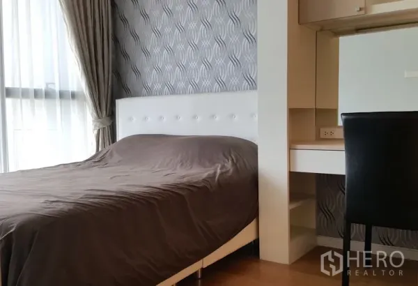 Condominium for rent in Sathon, Bangkok - Cozy bedroom with white tufted headboard, patterned wallpaper, built-in desk and cabinets, and a curtained window.