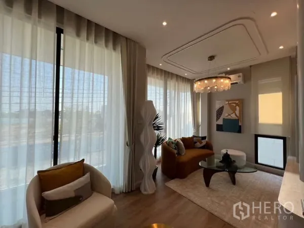 Detached House for sale in Saphan Sung, Bangkok - Sunlit living room with floor-to-ceiling curtains, chandelier, modern sofa and glass coffee table in a Saphan Sung Bangkok detached house.