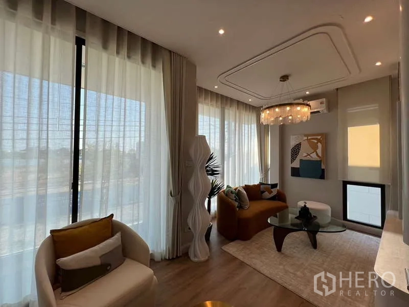 Detached House for sale in Saphan Sung, Bangkok - Sunlit living room with floor-to-ceiling curtains, chandelier, modern sofa and glass coffee table in a Saphan Sung Bangkok detached house.