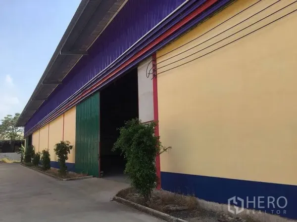 Warehouse for rent in Sai Noi, Nonthaburi - Colorful warehouse exterior with purple roof, yellow walls and a large green sliding door along a concrete driveway.