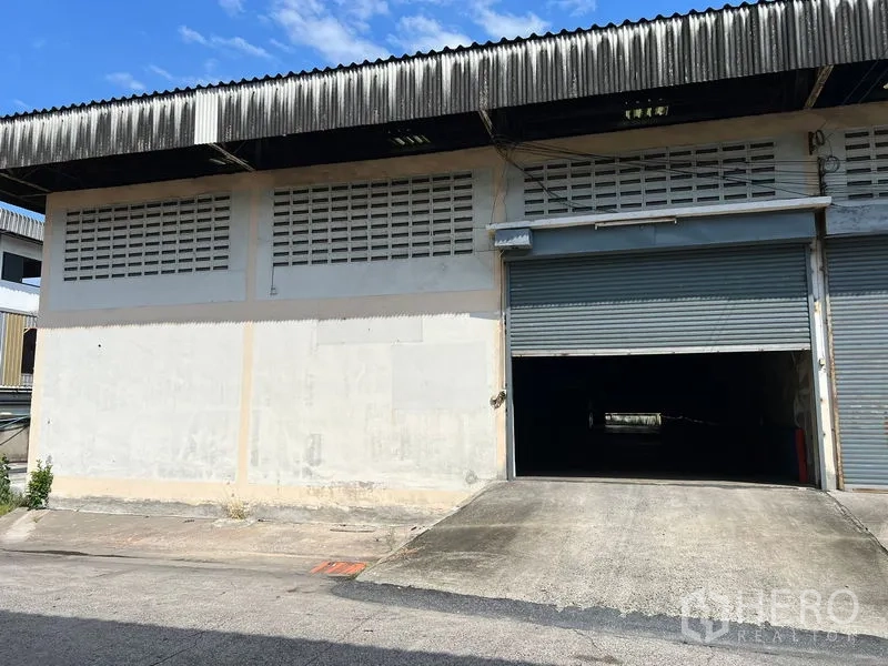 คลังสินค้า ให้เช่าในMueang Samut Prakan, Samut Prakan - ด้านหน้าโกดังมีประตูม้วนขนาดใหญ่ ทางลาดสำหรับขนถ่ายสินค้า และผนังบล็อกช่องลม