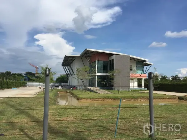 工厂 出租在Lam Luk Ka, Pathum Thani - Lam Luk Ka现代化工厂外立面，玻璃办公区、草坪、宽阔车道与水景，蓝天白云下。