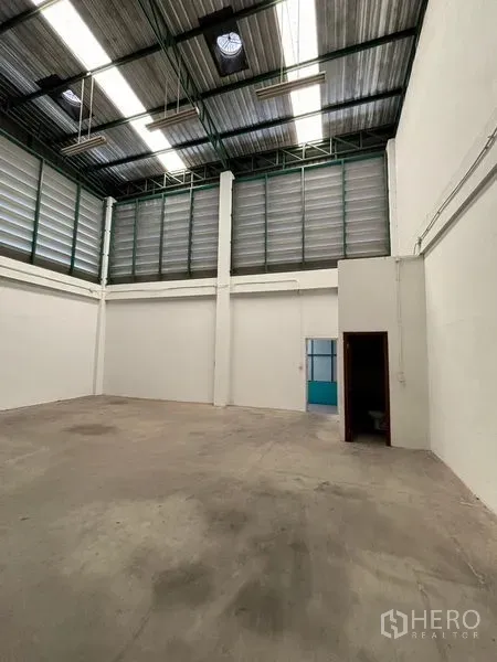 Warehouse for rent in Phra Samut Chedi, Samut Prakan - Open storage bay with tall ventilated walls, a blue side door and a small utility room.