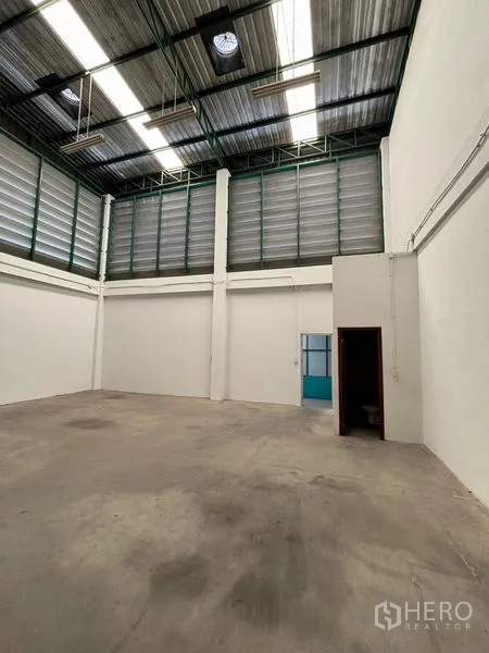 Warehouse for rent in Phra Samut Chedi, Samut Prakan - Open storage bay with tall ventilated walls, a blue side door and a small utility room.