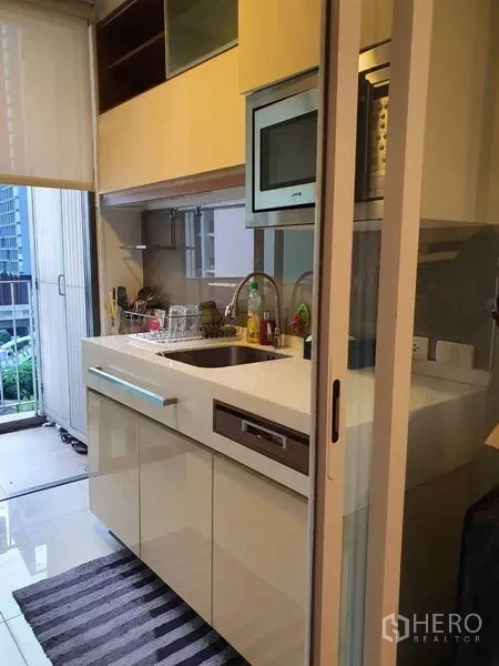 Condominium for sale in Bangkok - Modern kitchenette with sink, built-in microwave, glossy cabinets, and access to the balcony through a sliding door.