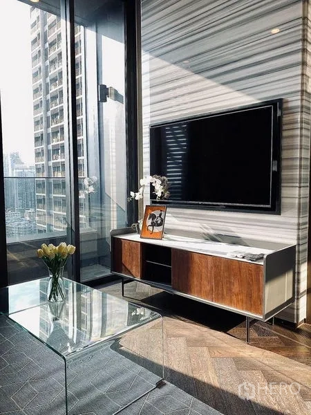 Condominium for rent in Khlong Toei, Bangkok - Living area with TV wall, glass coffee table, and sliding door to a balcony overlooking the city.