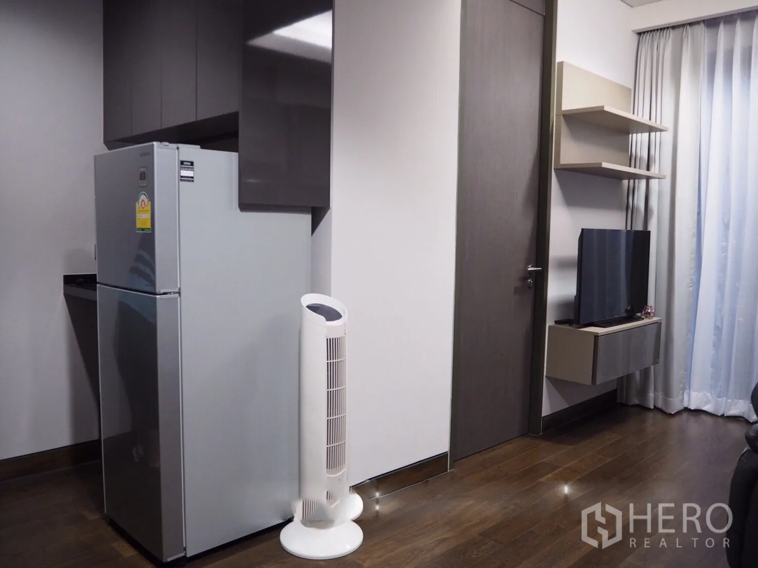 Condominium for rent in Khlong Toei, Bangkok - Entry corner with full-size refrigerator, tower fan and wall-mounted TV on wooden floor.