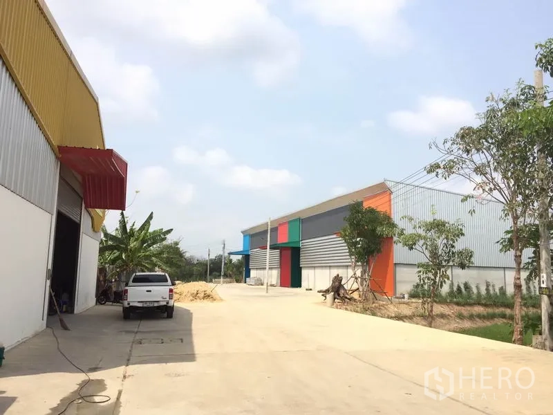 Warehouse for rent in Sai Noi, Nonthaburi - Wide driveway between modern warehouses with trees and colorful facades for easy truck access.