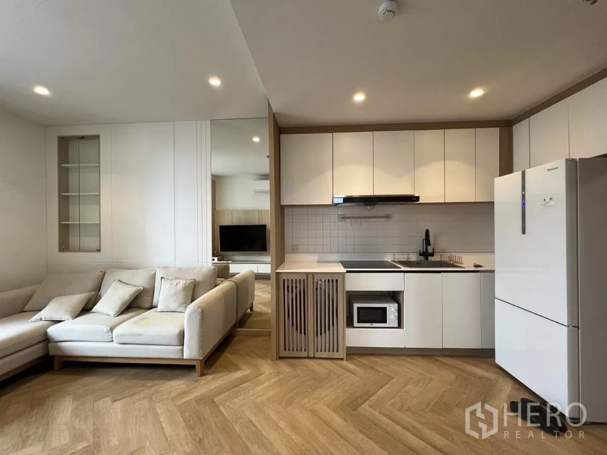 Condominium for rent or sale in Chatuchak, Bangkok - White one‑wall kitchen with cooktop, hood, microwave and large fridge beside a beige sectional sofa.