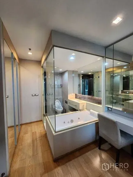 Condominium for sale in Ratchathewi, Bangkok - Glass-walled bathroom with soaking tub, mirrored vanity and a dressing table over warm wood flooring.
