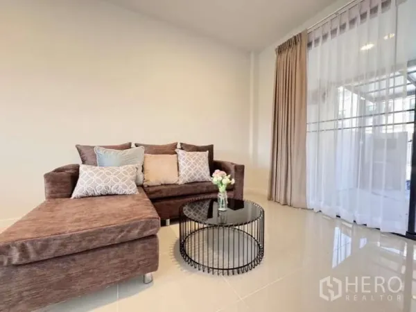 Townhouse for rent in Min Buri, Bangkok - Cozy living room featuring an L-shaped sofa, round glass coffee table and sliding glass door.