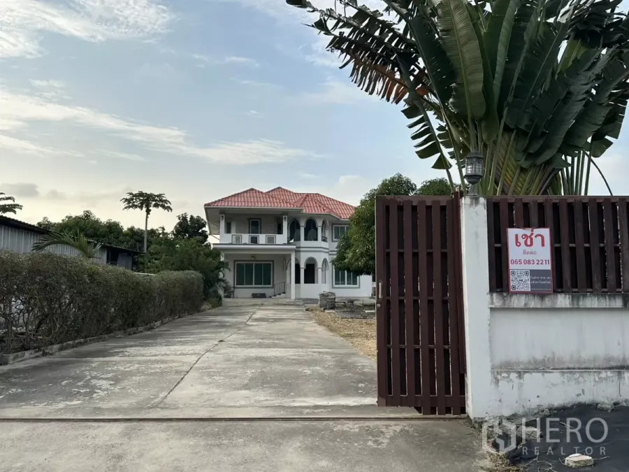 สำนักงานที่บ้าน ให้เช่า - ทางเข้ามีประตูรั้วและถนนคอนกรีตยาวสู่โฮมออฟฟิศสองชั้นสีขาวและลานกว้าง.